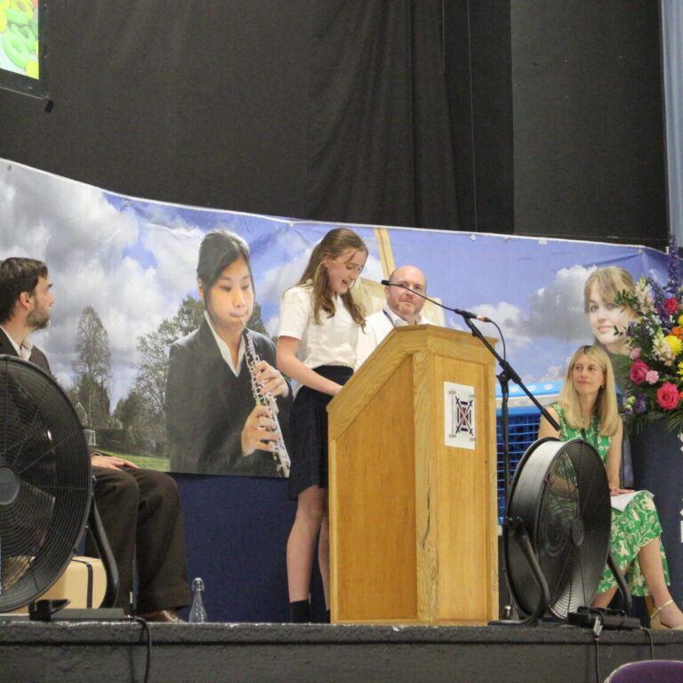 Students perform on a stage with a podium, banner reading 'Exeter School', and colorful flowers in the foreground.