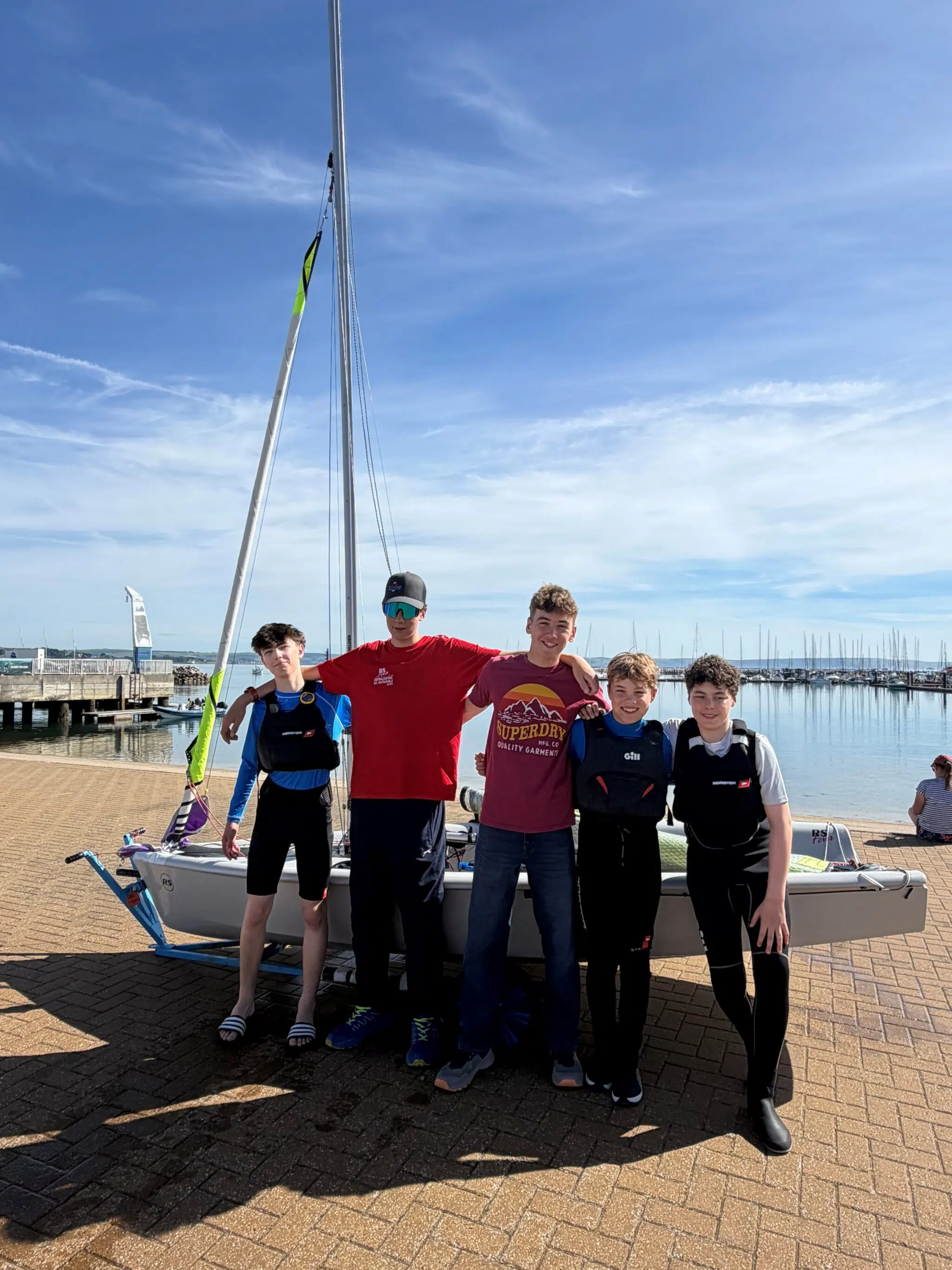 Group of six teenagers in life jackets posing in front of a small sailboat at a marina on a sunny day.