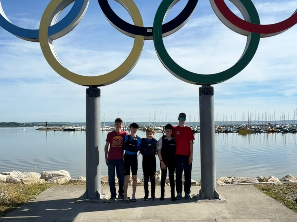 Six people posing under the large Olympic rings sculpture by a marina on a sunny day.