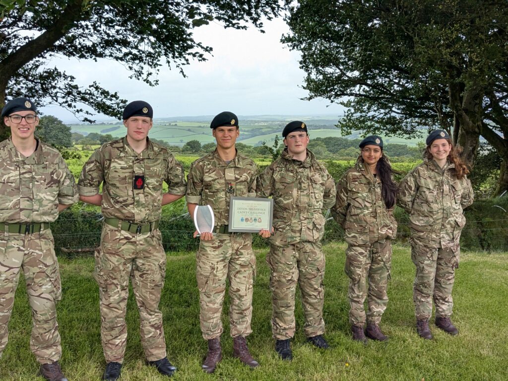 Six cadets in camouflage uniforms and berets stand in a row outdoors, holding a certificate and a trophy, with a rural landscape behind them.