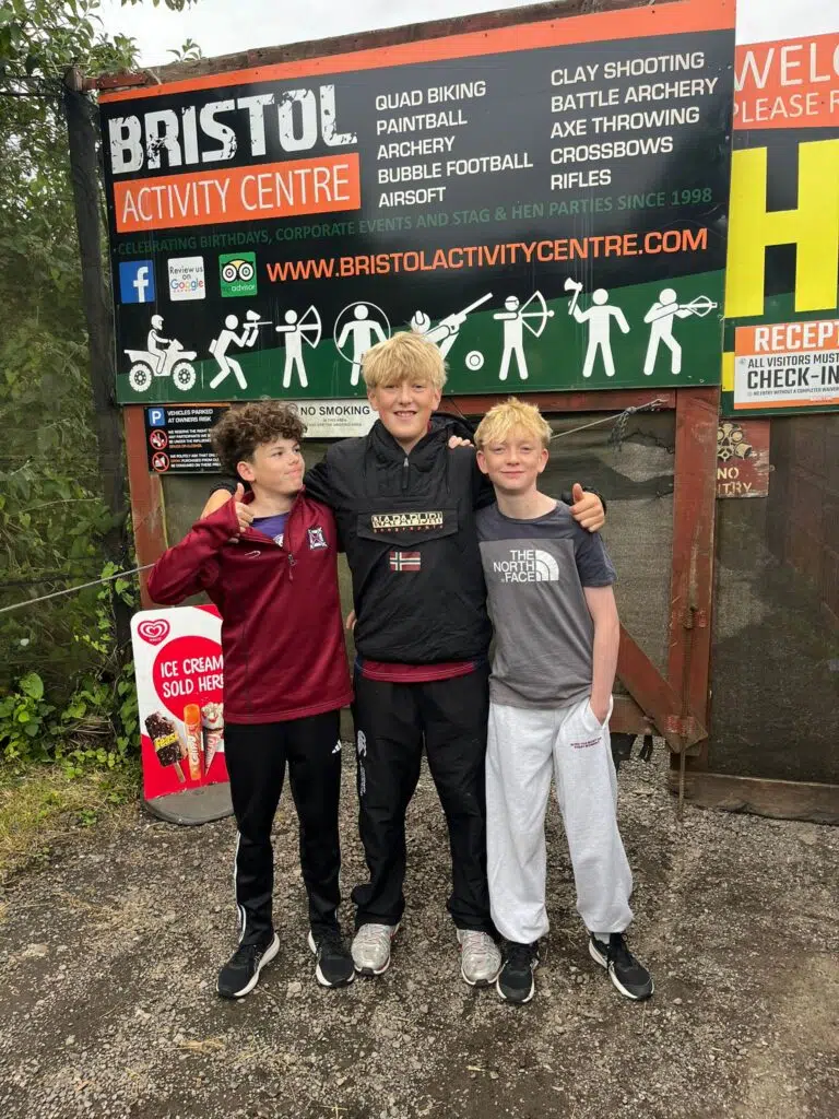 Three boys posing and smiling in front of a Bristol Activity Centre sign listing activities like quad biking and archery.