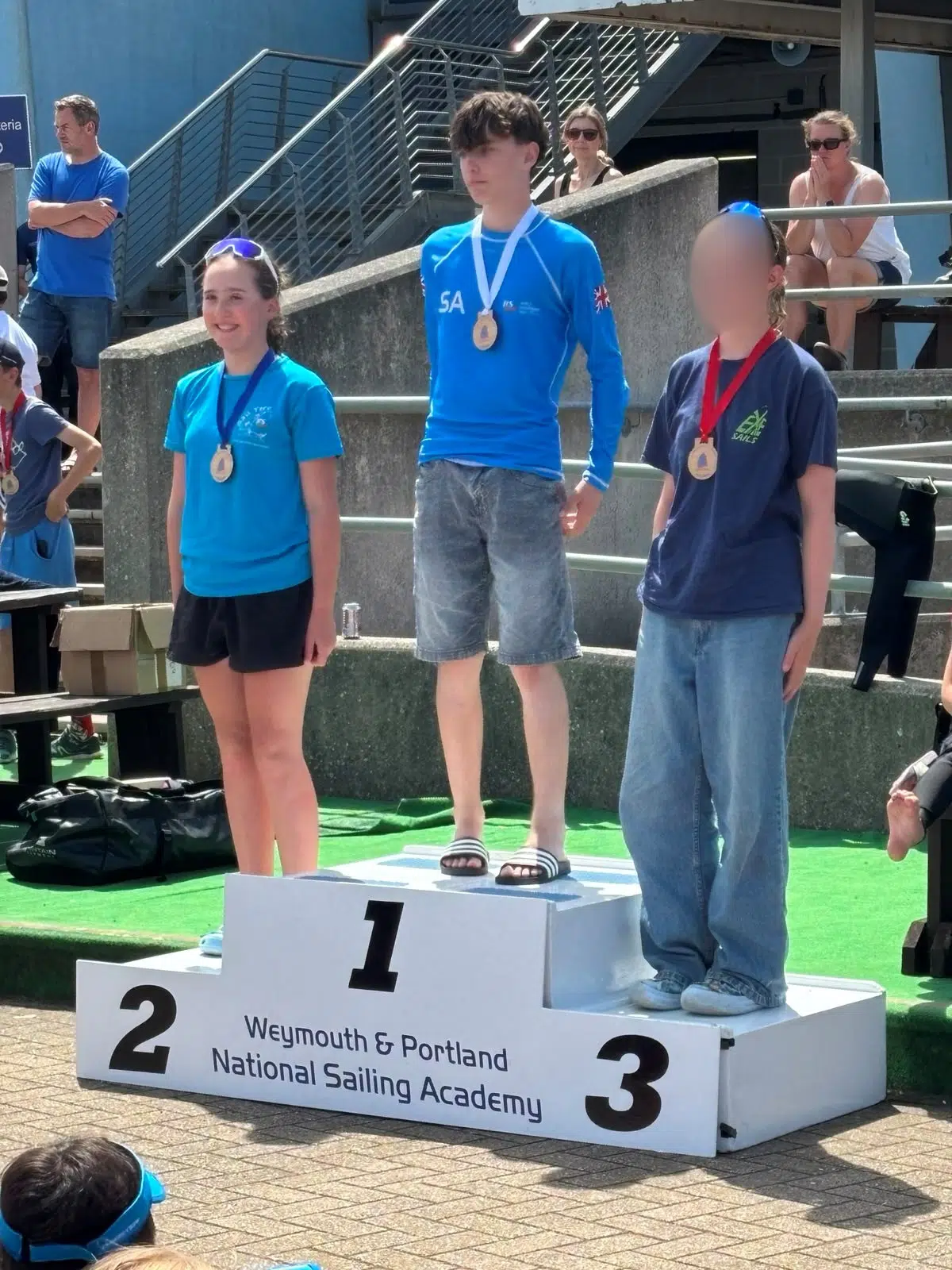 Three young competitors stand on a winners' podium during a sailing event, with the first-place center, second left, and third right, wearing medals and blue shirts; the podium reads Weymouth & Portland National Sailing Academy.