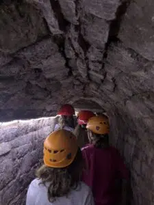Group of people wearing orange and yellow safety helmets walking through a narrow, rocky cave tunnel side-by-side.