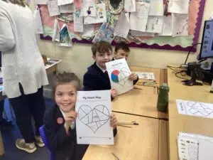 Three students in school uniforms sit at a classroom table, smiling and showing their 'My Heart Map' heart-shaped worksheets with colored sections.