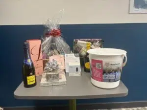 Table of gifts including a champagne bottle, wrapped baskets, and a white Hospice charity bucket on a gray table against a blue wall.