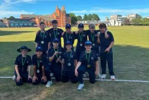 Group of young athletes posing on a grassy field with medals and a trophy, in matching dark uniforms, some kneeling and some standing with arms around each other.