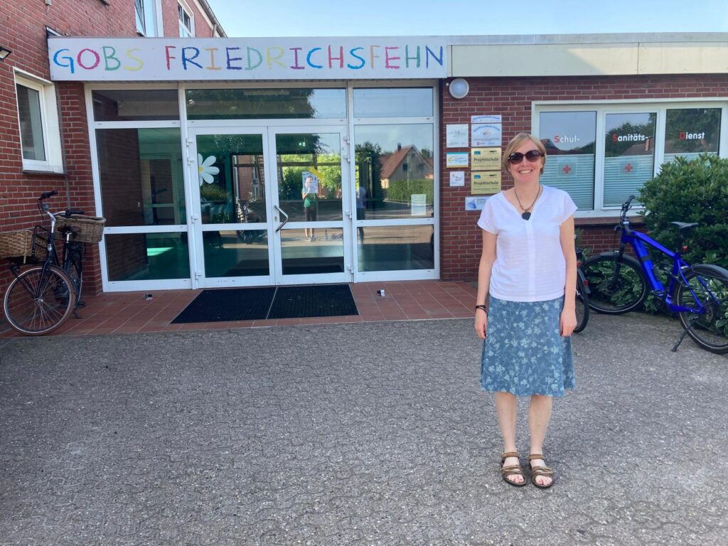 Smiling woman in a white top and blue skirt stands outside a brick building with a colorful banner above the glass doors, bikes on either side. - Alternative 1 for clarity: a woman posing in front of a school/community entrance with bicycles nearby.