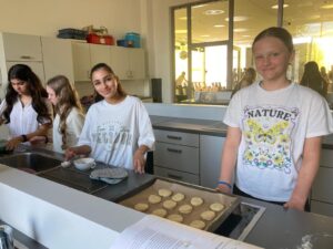 Group of young girls in a bright kitchen baking cookies at a counter, trays of dough rounds ready for baking visible in front of them