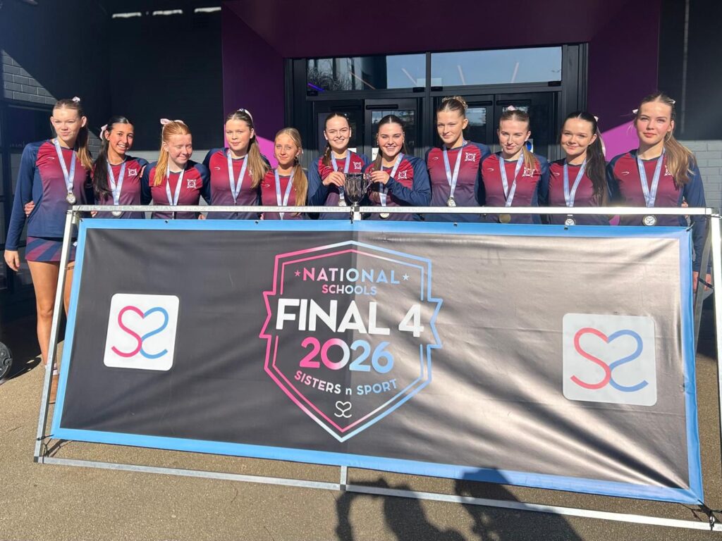 Group of young female athletes in matching maroon and navy uniforms pose with medals behind a banner that says National Schools Final 4 2026 — Sisters in Sport.