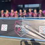 Group of young female athletes in matching maroon and navy uniforms pose with medals behind a banner that says National Schools Final 4 2026 — Sisters in Sport.