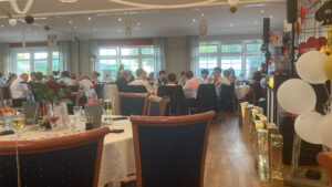 Busy banquet hall with groups at round tables, festive playing-card decorations, and balloons along a display of gift boxes on the right.