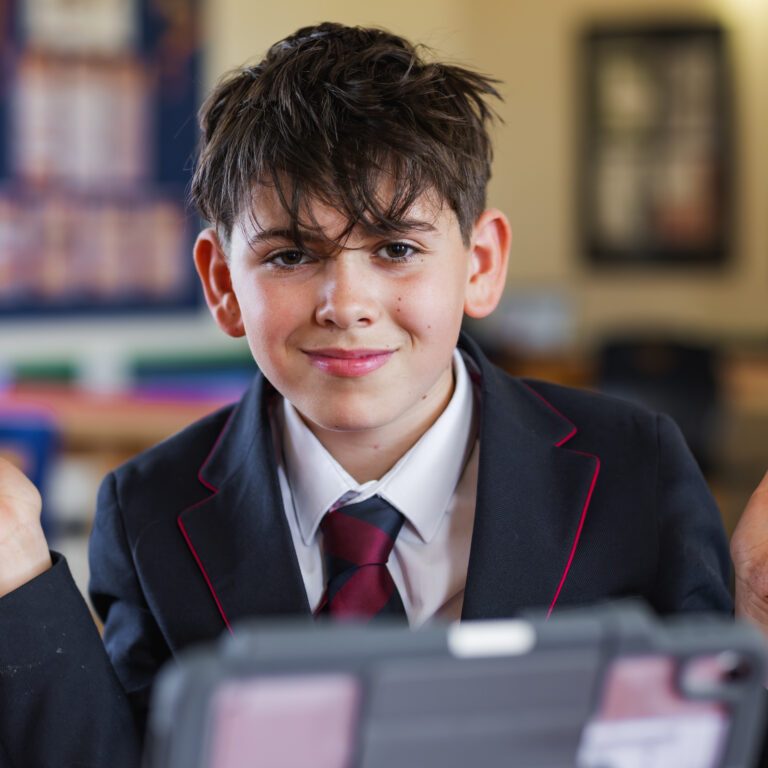 Smiling student in a navy blazer and red tie raises his hands in a classroom, with a tablet in front.