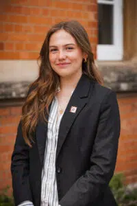 Portrait of a smiling young woman with long brown hair, wearing a black blazer and striped blouse, standing outside against a red brick wall.