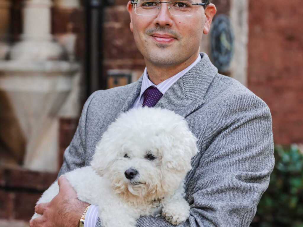 Man in a gray blazer holding a small white fluffy dog outdoors, brick building in the background.