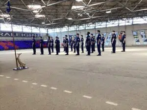 Line of uniformed cadets standing at attention in a large indoor hall, a flag on a stand in the foreground.