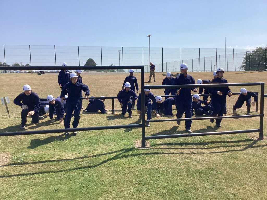 Group of people in navy uniforms and white helmets sprinting through a low metal fence obstacle on a sunny field, with a chain-link fence in the background.