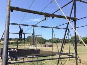 Person wearing a white safety helmet climbs a metal obstacle frame outdoors on a clear day with blue sky behind it.