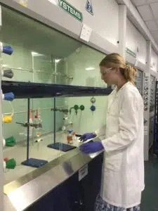 Female scientist in a lab coat and safety goggles standing at a fume hood, handling glassware and bottles on the bench.