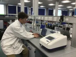 Lab technician in a white coat and safety goggles typing on a laptop at a lab bench.