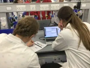 Two students in lab coats and safety goggles study data on a laptop at a lab bench, with charts on paper nearby.