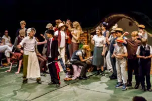 Group of students in vintage costumes performing on a theater stage, some standing and others kneeling, with a rustic backdrop and audience visible on the left.