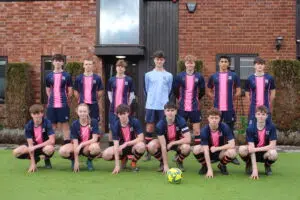 Youth soccer team in pink-and-navy kits pose for a group photo on artificial turf, a ball at their feet in front of a brick building.