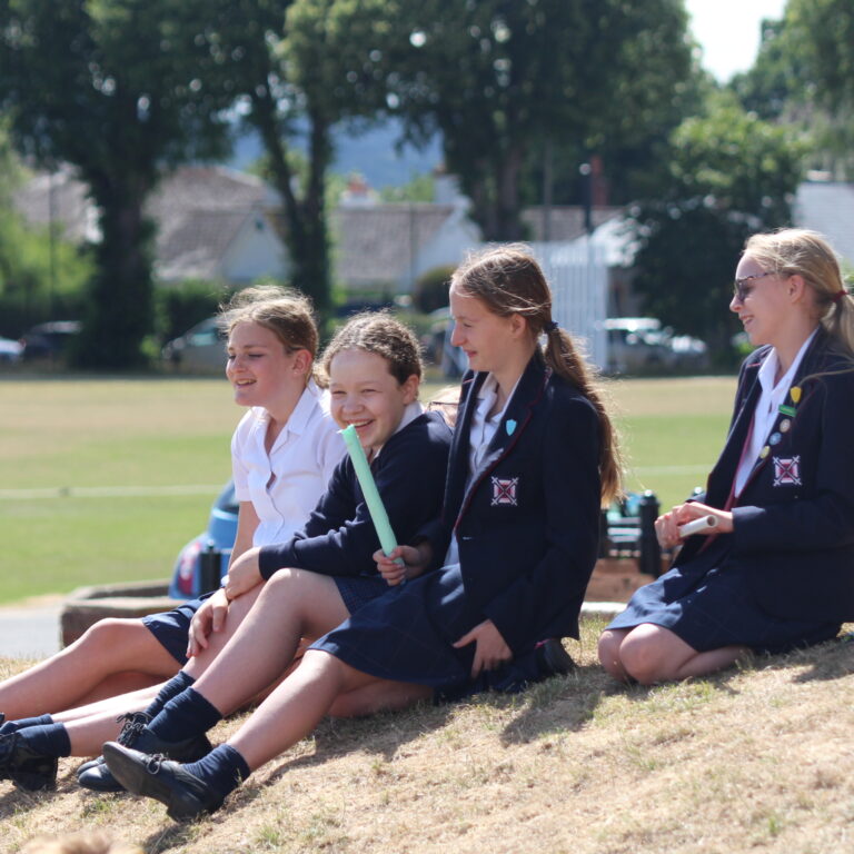 Four schoolgirls in navy uniforms sit on a grassy bank outdoors, smiling and chatting on a sunny day.