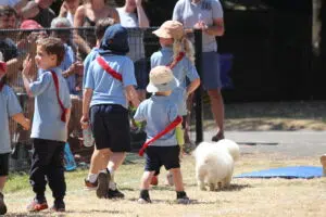 Group of children in light-blue shirts with red sashes standing near a fence at an outdoor event, with a small white dog in the foreground and spectators behind the fence.