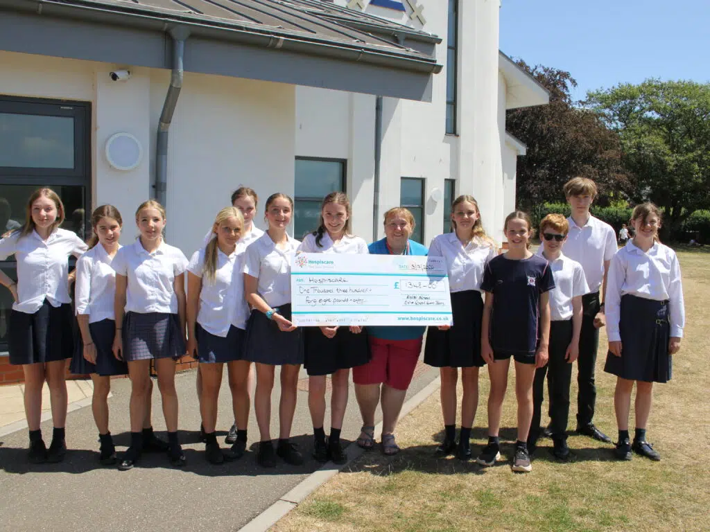 Group of students in uniform posing outside a building, holding a large charity check to Hospiscare.