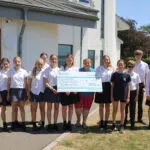 Group of students in uniform posing outside a building, holding a large charity check to Hospiscare.