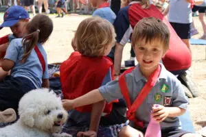 Children in gray shirts with red sashes gather on a blanket at an outdoor event; a fluffy white dog sits in front of them.