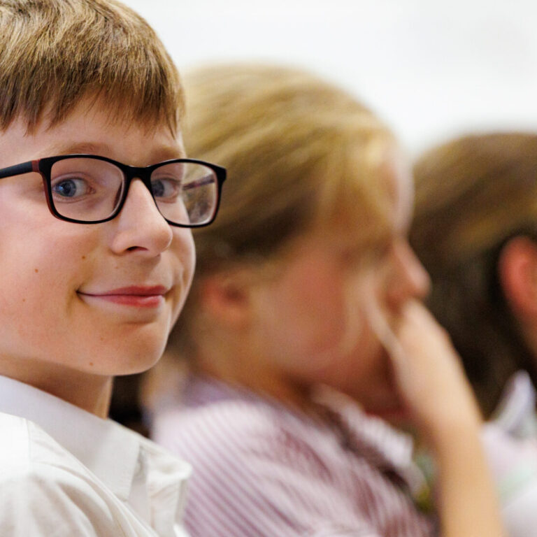Smiling boy with glasses in a white shirt, seated among classmates in a classroom.