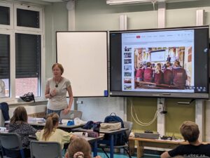 Teacher standing at front of classroom as students sit at desks; a large display shows a photo of kids in red uniforms presenting.