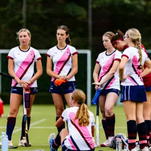 Group of female field hockey players standing on a green turf, wearing white jerseys with pink diagonal stripes and holding sticks.