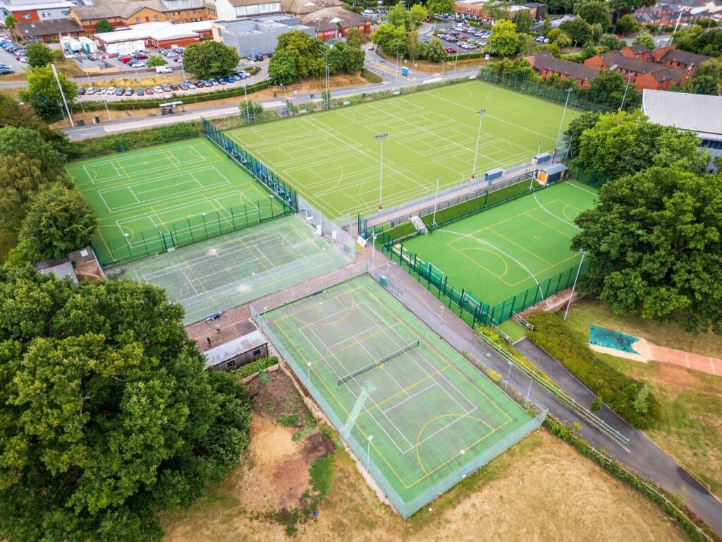 Aerial view of a multi-sport complex with fenced tennis courts and large green fields surrounded by trees and buildings nearby.