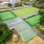Aerial view of a multi-sport complex with fenced tennis courts and large green fields surrounded by trees and buildings nearby.