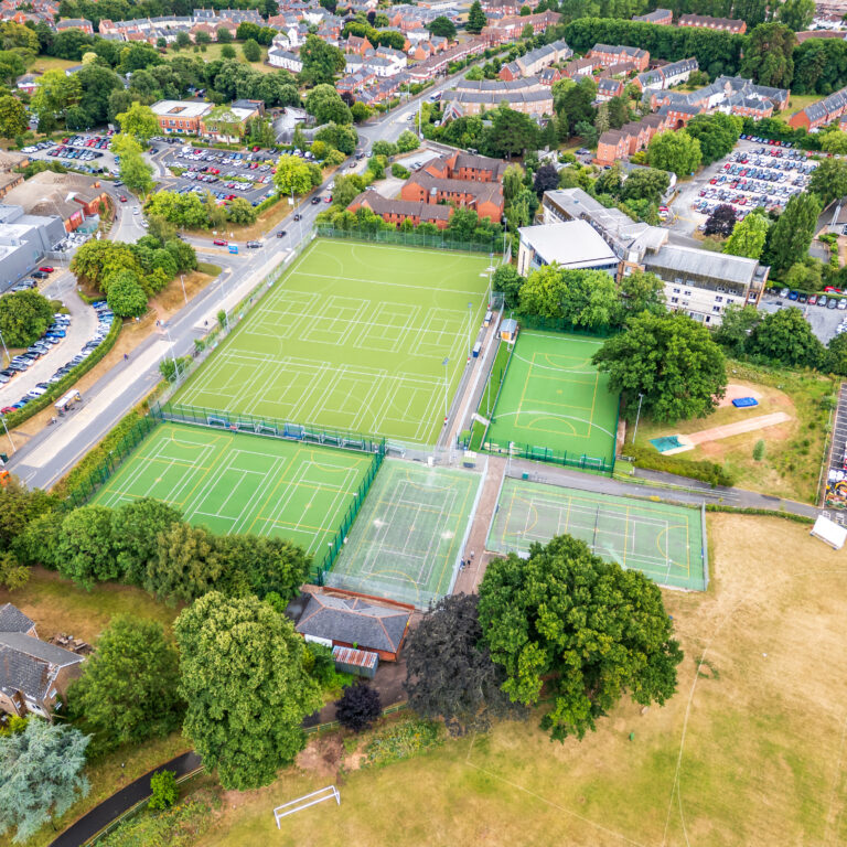 Aerial view of a sports complex with multiple green turf fields fenced in, surrounded by roads, trees, and a nearby residential area and parking lots.