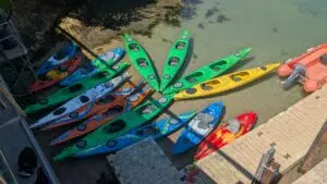 Cluster of bright green, yellow, orange, blue, and red kayaks tied along a wooden dock by clear shallow water, viewed from above.