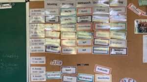 Weekly class timetable on a corkboard, with German days (Montag–Freitag) and subjects pinned in colorful cards: Englisch, Mathematik, Deutsch, etc.