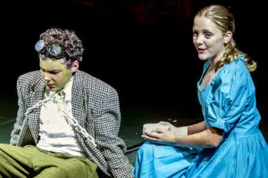 Two actors sit on a dark stage: a man in a houndstooth jacket with goggles and a white ruffled shirt, looking down, while a woman in a bright blue dress holds a white cup and smiles slightly at him.