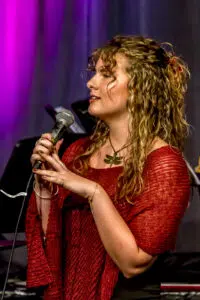 Singer with curly hair wearing a red knit top, holding a microphone on stage under purple lighting.
