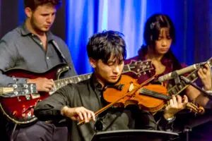 Violinist in black plays violin while two guitarists perform beside him on a blue-lit stage.