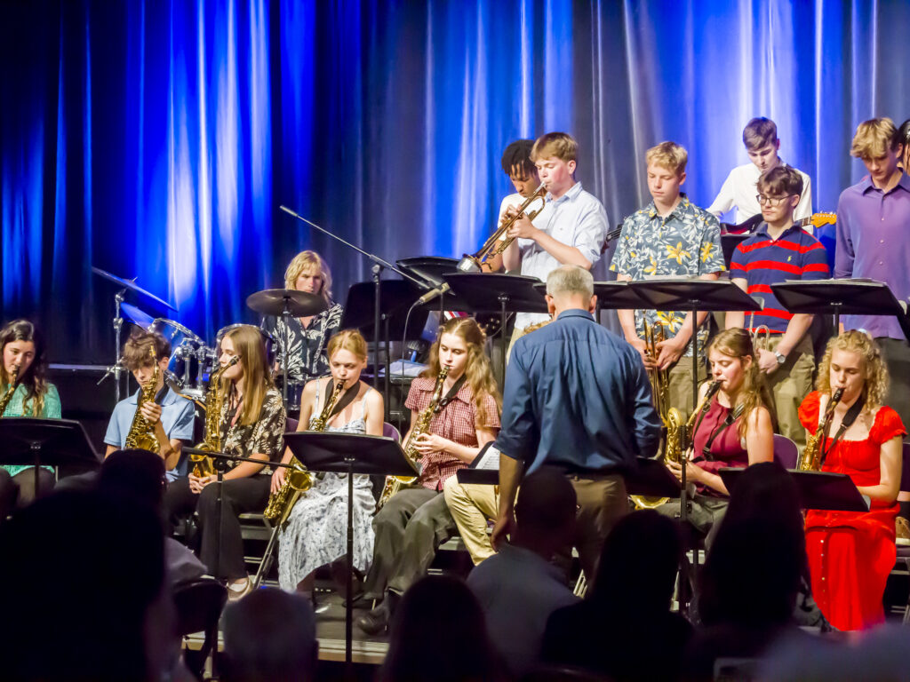 Large group of young musicians performing on a stage with brass and woodwinds under blue lights; conductor in front.