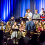 Large group of young musicians performing on a stage with brass and woodwinds under blue lights; conductor in front.