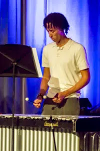 Young man in a white polo shirt playing a marimba on stage, holding mallets and looking at the instrument in front of blue curtains.