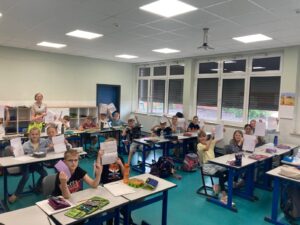 Smiling teacher stands at back of classroom as students at desks raise their completed worksheets for the photo.