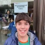 Smiling teenage boy in teal shirt and blue jacket stands outside a glass door with a sign behind him reading ‘NCO orchestral weekends Midlands’