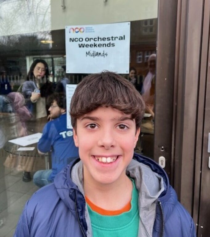 Smiling teenage boy in teal shirt and blue jacket stands outside a glass door with a sign behind him reading ‘NCO orchestral weekends Midlands’