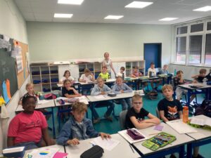 Classroom full of young students at desks, smiling, with a teacher at the back and school supplies on shelves along the wall.