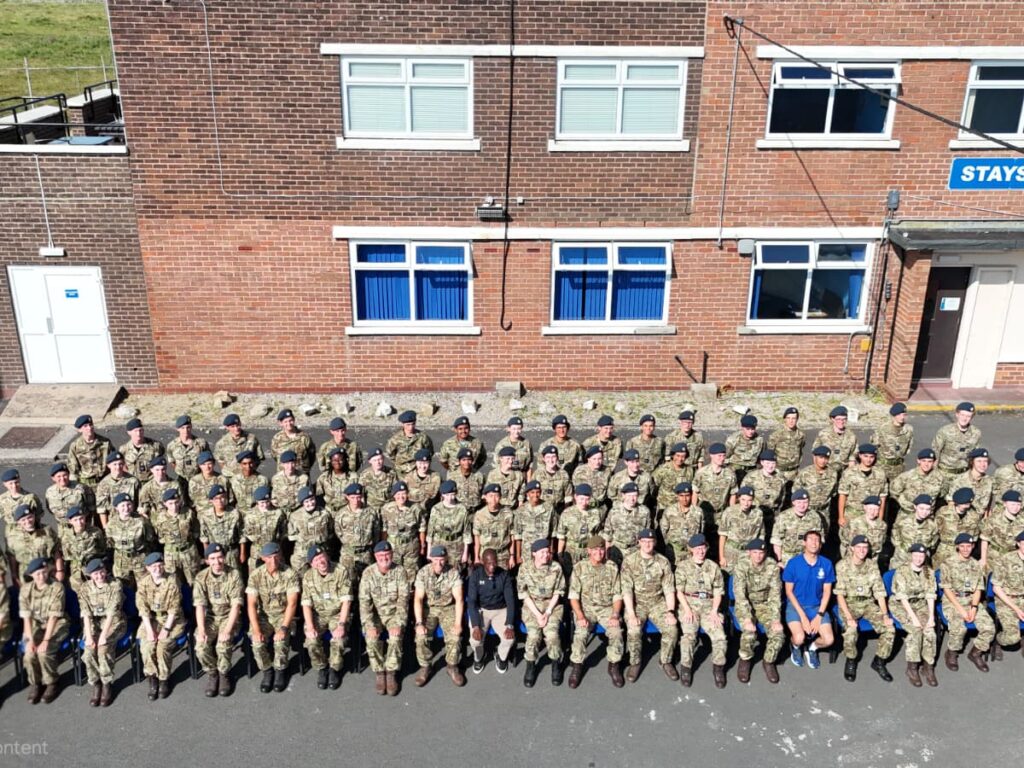 Group of soldiers in camouflage uniforms posing outdoors in front of a brick building with blue windows and a sign nearby.